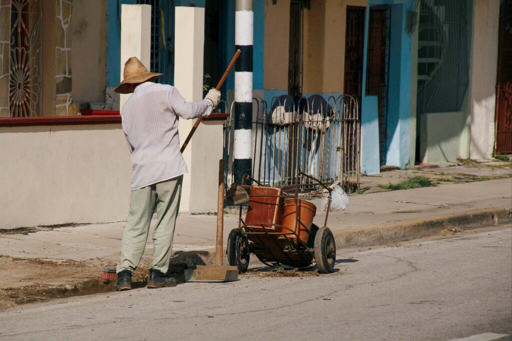 A solitary worker sweeps a sidewalk in an urban environment with a handcart and broom.